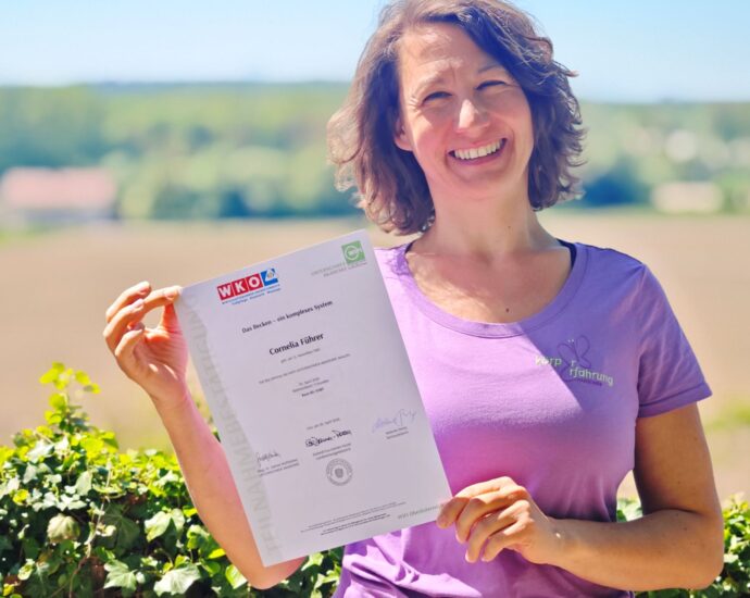 Frau im violetten Körpererfahrung-Shirt hält im Freien ein WIFI-Zertifikat zum Seminar „Das Becken – ein komplexes System“ in die Kamera.